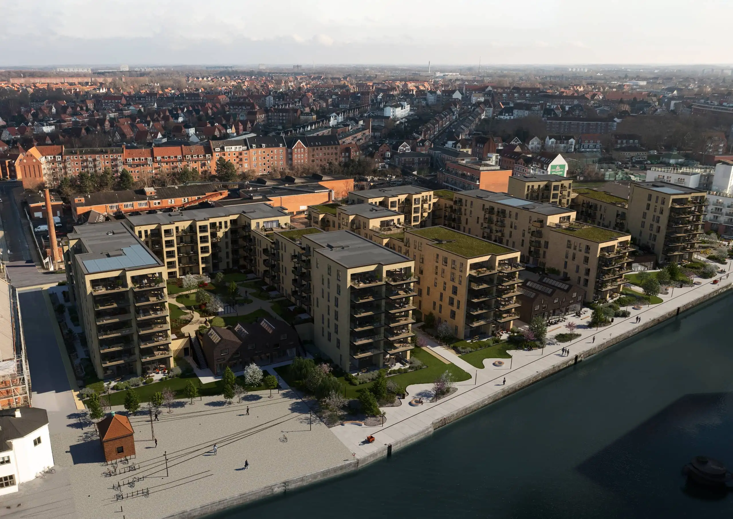 Luftfoto af moderne lejlighedskomplekser med grønne tage og anlagte gårde ved en havnefront, omgivet af ældre murstensbygninger og boligområder i baggrunden.