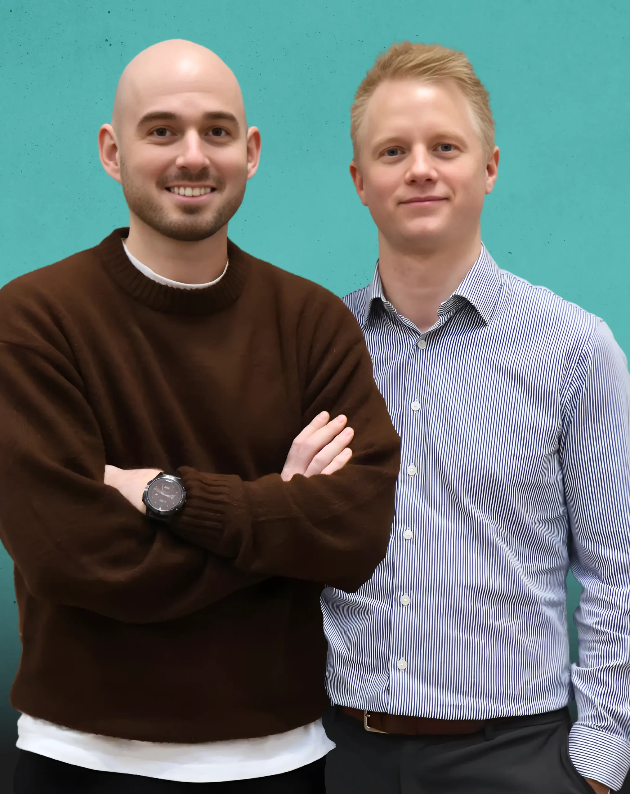 To mænd står side om side foran en blågrøn baggrund. Manden til venstre er skaldet, iført en brun sweater og et sort ur, med armene over kors. Manden til højre har lyst hår og er iført en stribet skjorte med knapper.