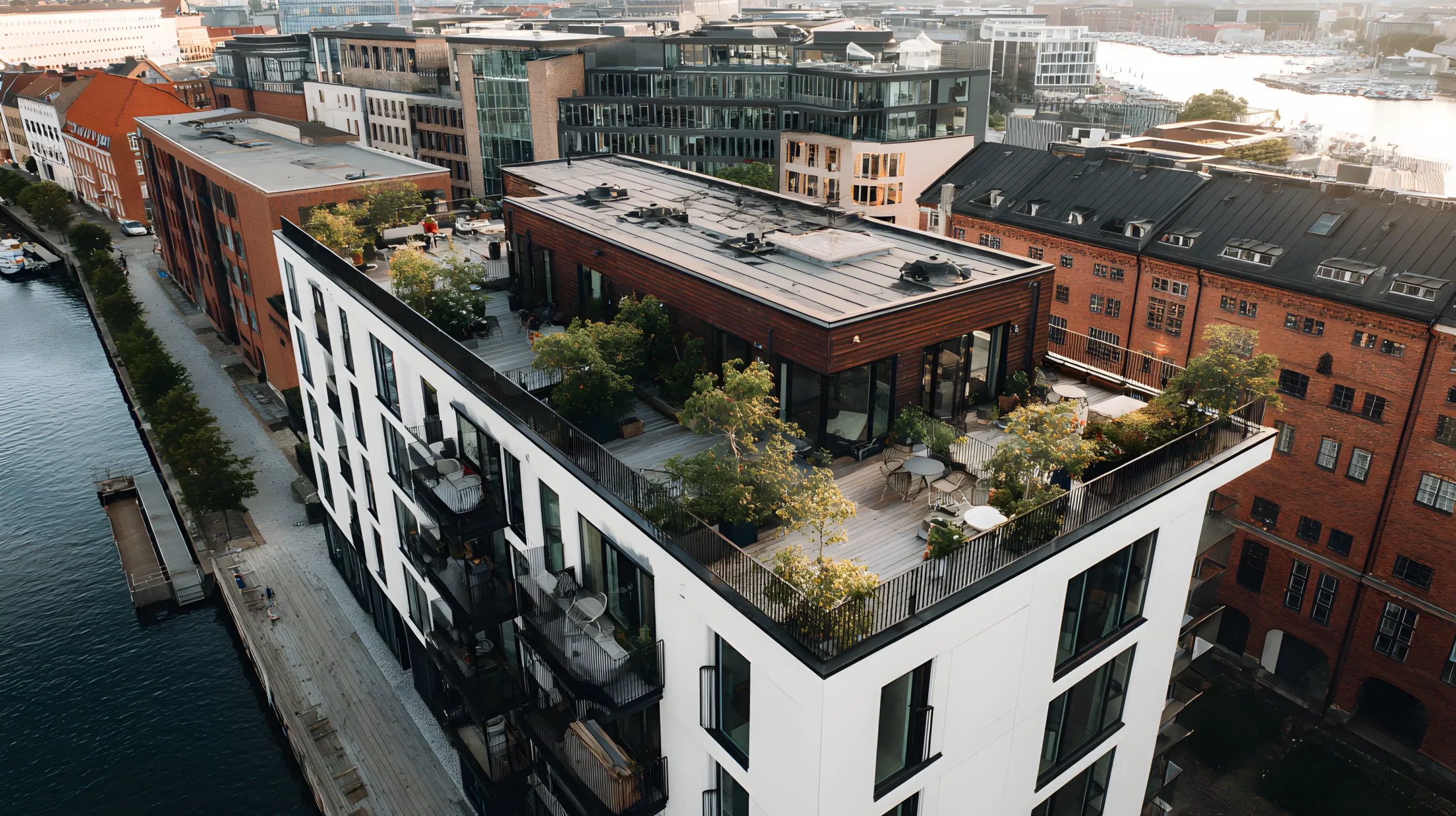 Et moderne hvidt lejlighedskompleks med en tagterrasse fyldt med grønt, omgivet af ældre murstensbygninger og beliggende ved siden af en kanal i et urbant miljø.