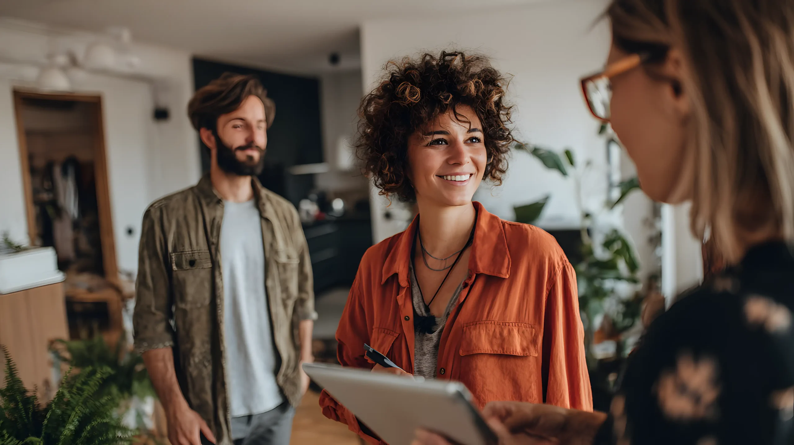 Tre mennesker taler sammen i en lys, moderne stue. En kvinde med krøllet hår og orange skjorte smiler til en anden kvinde, der holder et clipboard, mens en mand står i baggrunden og følger med i samtalen.