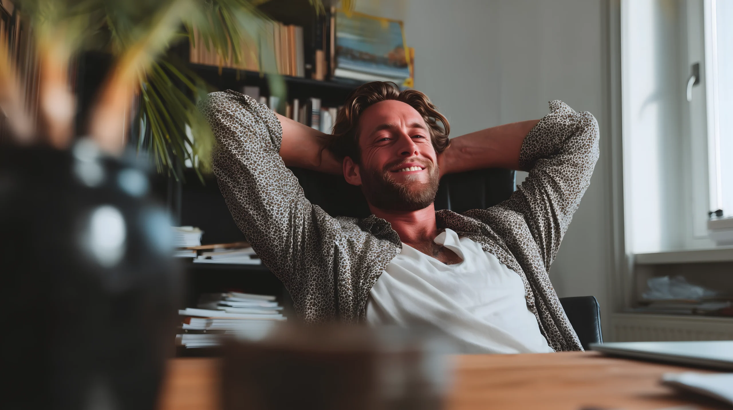 En mand smiler og læner sig tilbage i en stol med hænderne bag hovedet i et afslappet kontormiljø med bogreoler og et vindue i baggrunden.